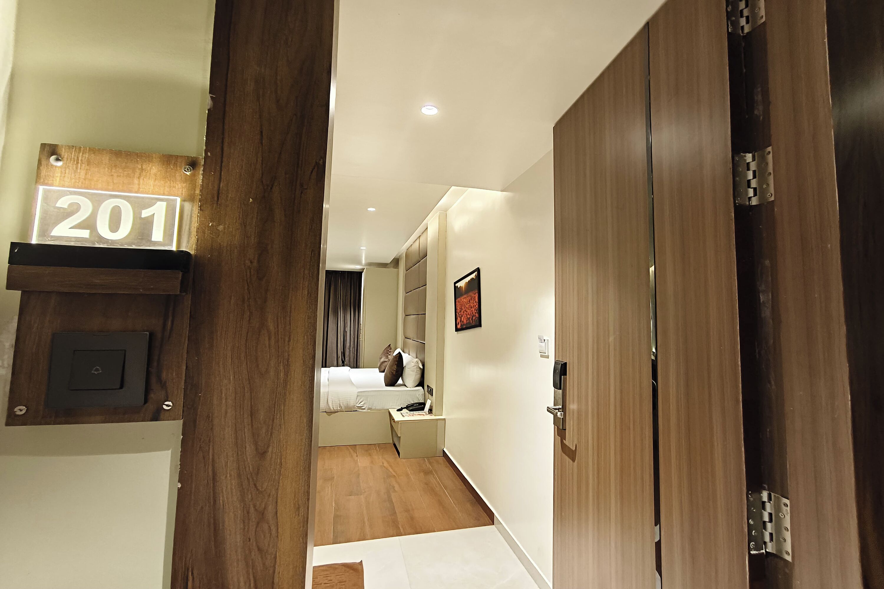 A hotel room with a wooden door, a number sign '201', a bed with white bedding, a bedside table, and a small desk with a chair.