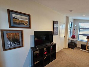 A living room with a flat-screen TV on a black stand, framed artwork on the walls, a small table with a vase, and a door with a 'home' sign. - Cosy Queenstown Studio for 2 (Queenstown)