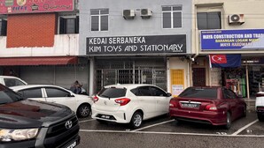 A parking lot with several cars, a building with a sign for 'Kedai Serbaena Kim Toys and Stationary', and another sign for 'Maya Construction and Trade'.