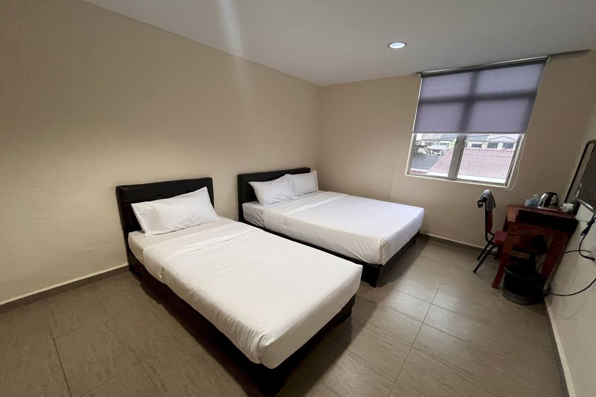 A hotel room with two single beds, a desk with a chair, and a window with a view of buildings.