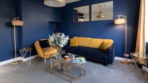 Ein Wohnzimmer mit blauer Wand, einem gelben Sessel, einem blauen Sofa, einem Glastisch und einer Pendelleuchte.