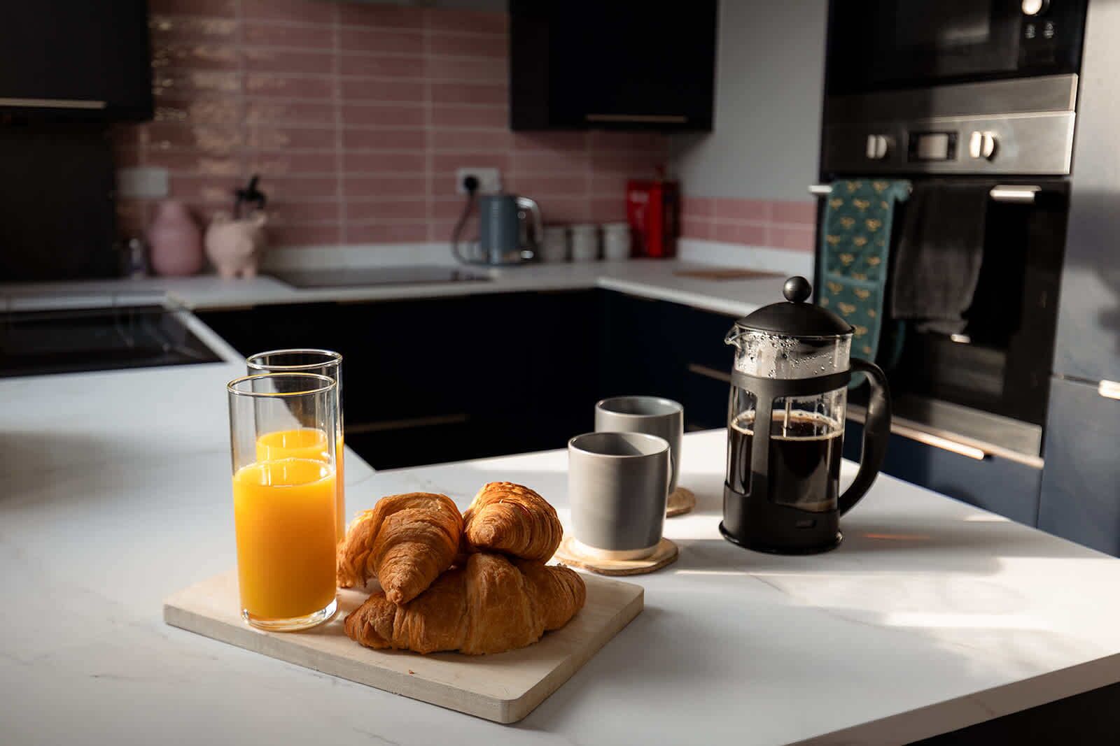 A kitchen counter with a glass of orange juice, two croissants, two cups, and a coffee press.