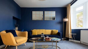 Ein Wohnzimmer mit blauer Wand, einem gelben Sofa, einem gelben Sessel, einem Glastisch mit Kaffeekanne und einer Stehlampe.