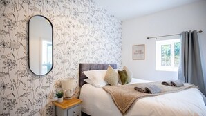 A bedroom with a floral wallpaper, a bed with white linens, a bedside table with a lamp, and a window with curtains.