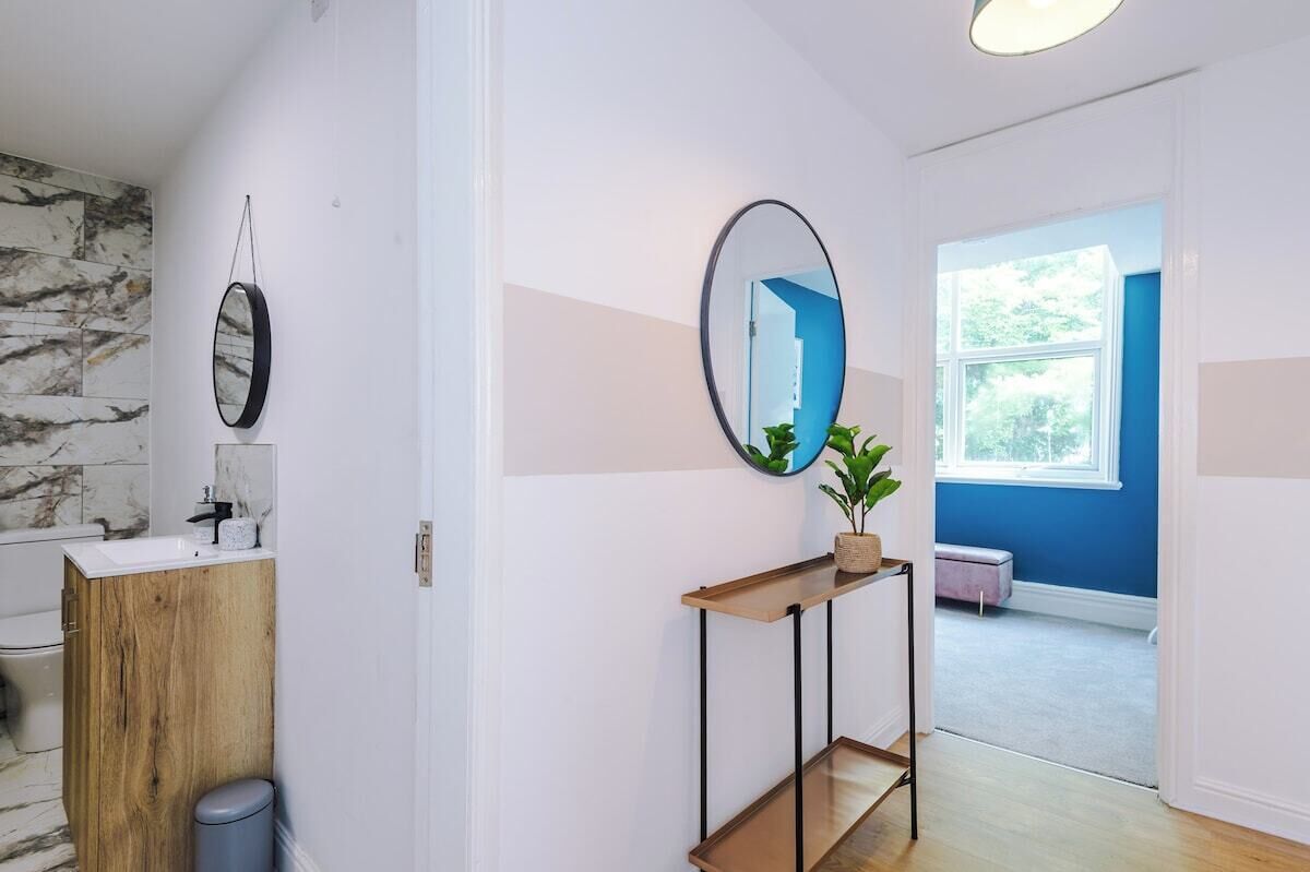A modern bathroom with a marble backsplash, a wooden vanity, a round mirror, and a potted plant on a console table.