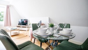 A dining area with a glass table set for four, surrounded by green upholstered chairs. A television is visible in the background, and the room has a modern, minimalist design. - The Anchorage Apartments - No.6 (Conwy Principal Area)