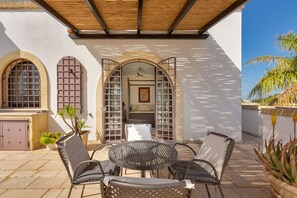 Une terrasse extérieure avec une table ronde et quatre chaises, un plafond en bois et un mur blanc agrémenté de fenêtres en arc de cercle.
