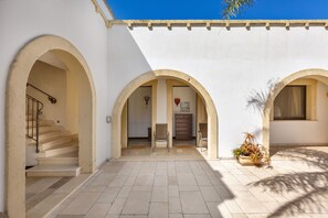 Une cour intérieure avec trois arcades, un escalier, une chaise en bois, une commode et un pot de fleurs.
