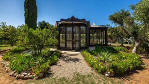 A conservatory with a glass door, surrounded by a garden with various plants and a tall tree. - 4015 Masseria Scilla by Barbarhouse (Carovigno)