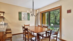 A dining area with a wooden table and chairs, a chandelier, a window with curtains, and a view of greenery outside.
