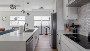 A modern kitchen with white cabinetry, a stainless steel refrigerator, and a black countertop. There is a window with blinds, a bar stool, and a hanging light fixture.
