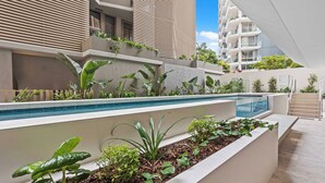 Un área de piscina moderna al aire libre con un jardín integrado y diversas plantas, una piscina azul cristalina y un edificio contemporáneo al fondo.