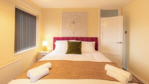 Une chambre d’hôtel comprenant un lit, une table de chevet, une lampe, une fenêtre avec des stores, une porte et un mur orné d’un tableau.