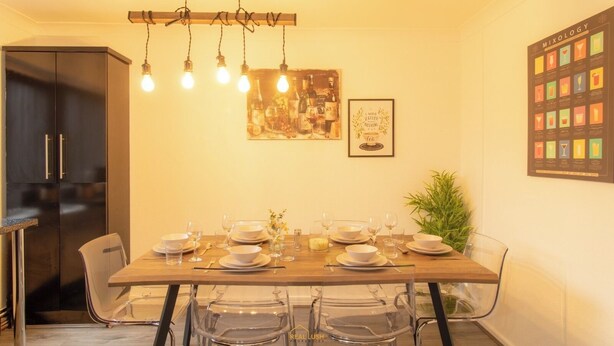 Un coin repas avec une table en bois dressée pour quatre personnes, des chaises en verre transparent, un luminaire suspendu, une œuvre d’art encadrée et un tableau illustrant différentes recettes de cocktails accroché au mur.
