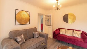 Un salon avec un canapé gris, un canapé rouge et un lustre. On y trouve deux éléments de décoration encadrés accrochés au mur, ainsi qu’une porte ornée d’un vitrail.