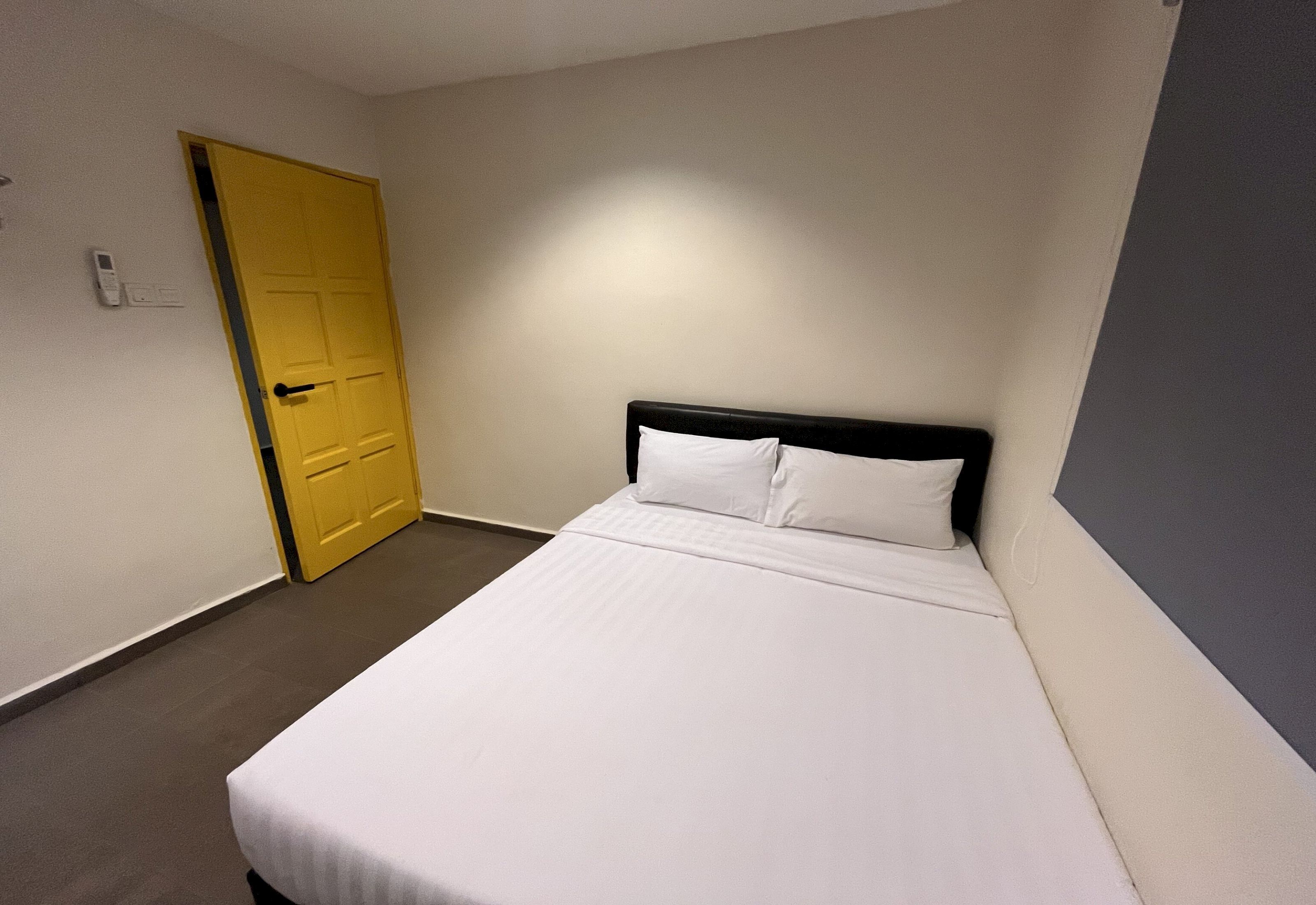 Un lit simple avec une literie blanche, dans une pièce dont la porte est jaune et les murs de couleur claire.