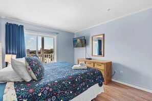 Relax in this cozy room with a large bed, TV, and dresser, featuring a floral-patterned duvet and a window with a view of the outdoors.