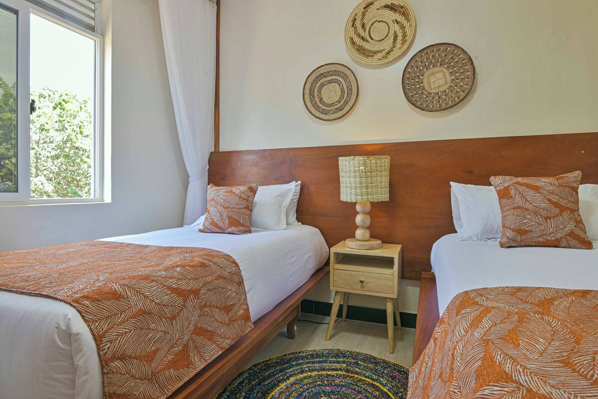 A hotel room with two beds, a wooden headboard, a bedside table with a lamp, and decorative wall hangings.
