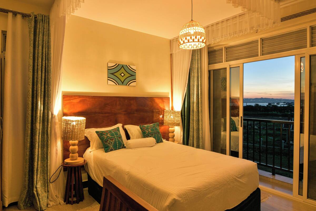 A bedroom with a large bed, two bedside lamps, a view of the cityscape through a sliding glass door, and a decorative ceiling light.
