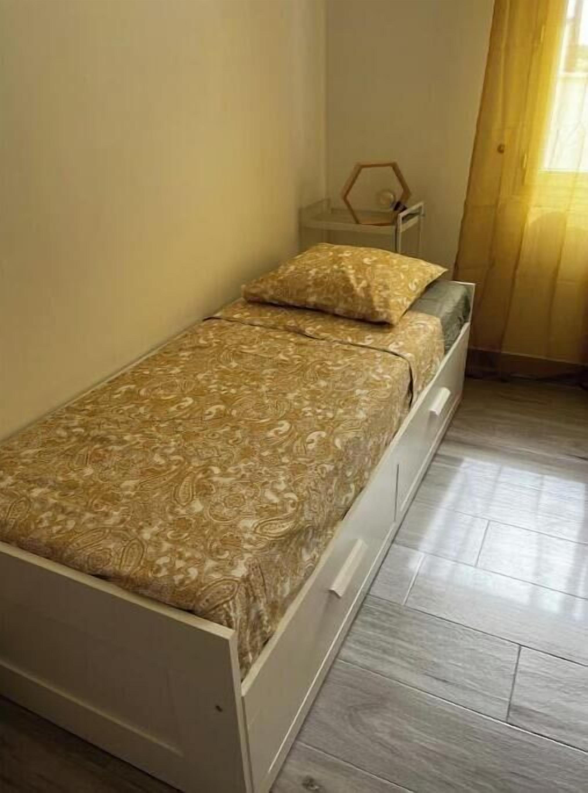 A small, single-bed room with a patterned bedspread, a bedside table, and a window with curtains.