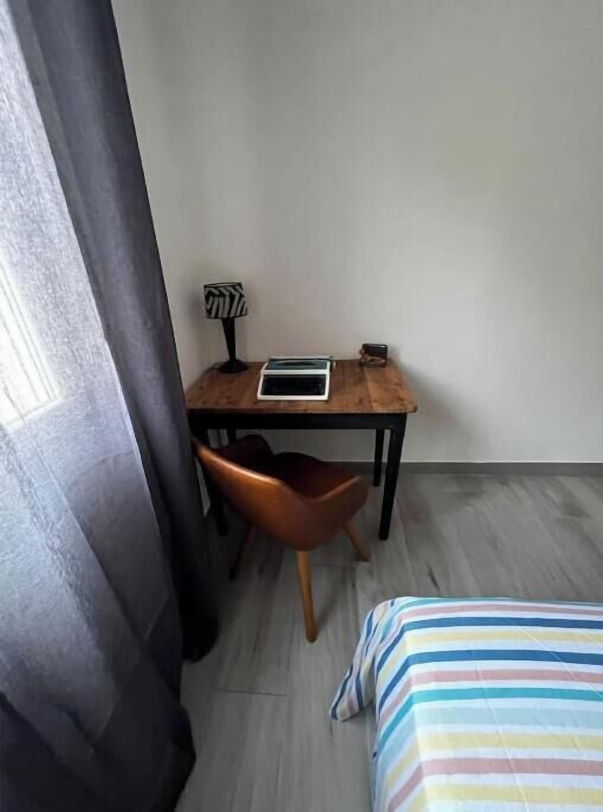 A small, neatly arranged room with a wooden desk, a chair, a bed with striped bedding, and a window with curtains.