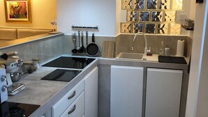 Une cuisine compacte équipée d’une cuisinière intégrée, d’un évier et d’un lave-vaisselle. Un étagère fixée au mur permet de ranger les ustensiles, et un revêtement mural décoratif en carrelage complète l’ensemble.