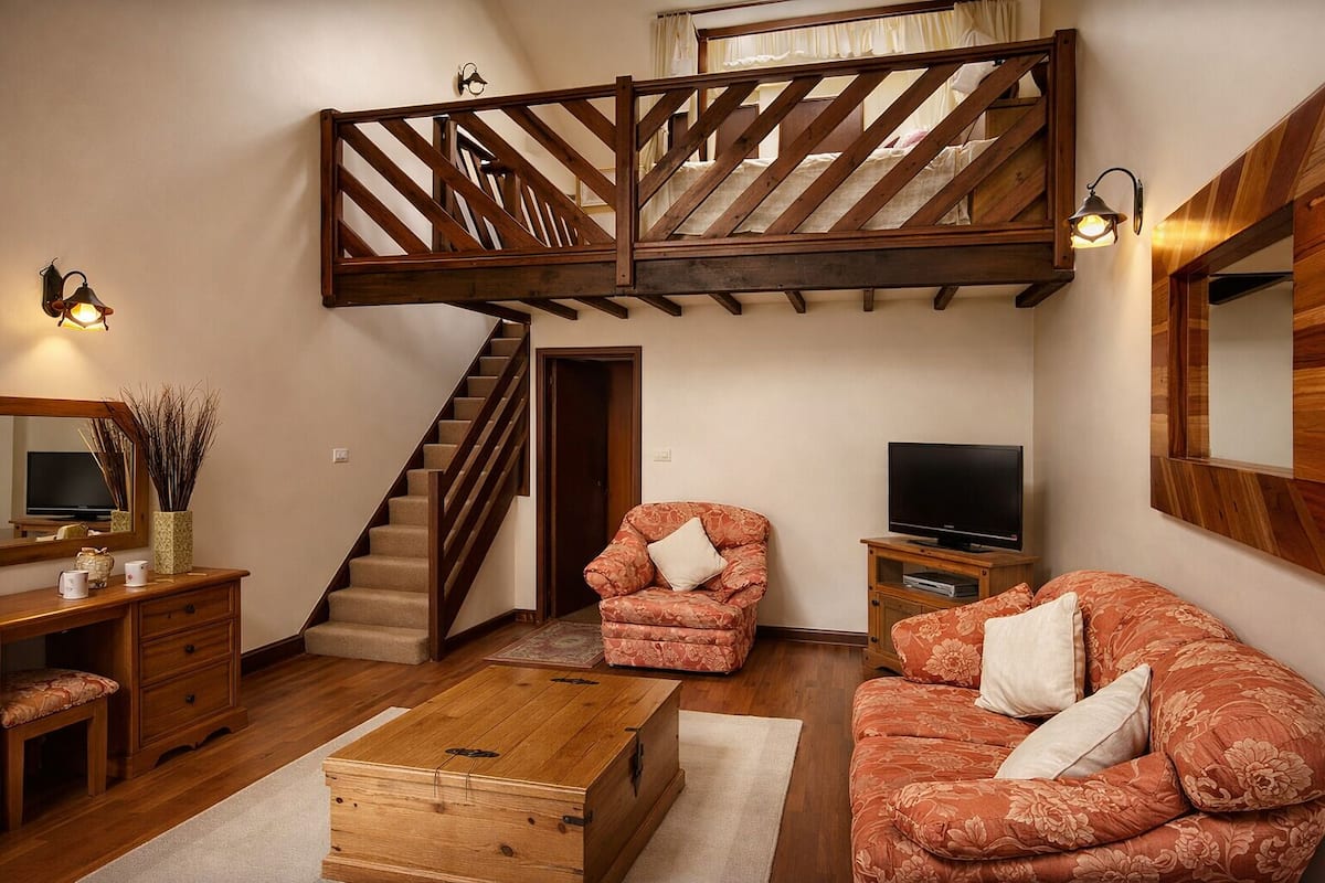A loft-style living room with a wooden staircase, a television, two floral-patterned sofas, a wooden coffee table, and a desk with a mirror and decorative items.