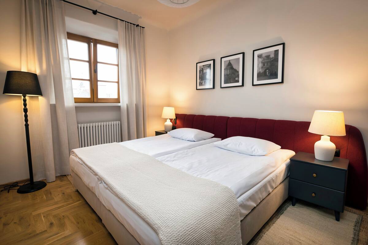 Un lit double avec du linge de lit blanc, une tête de lit rouge et deux tables de chevet avec des lampes. Trois tableaux encadrés ornent le mur.