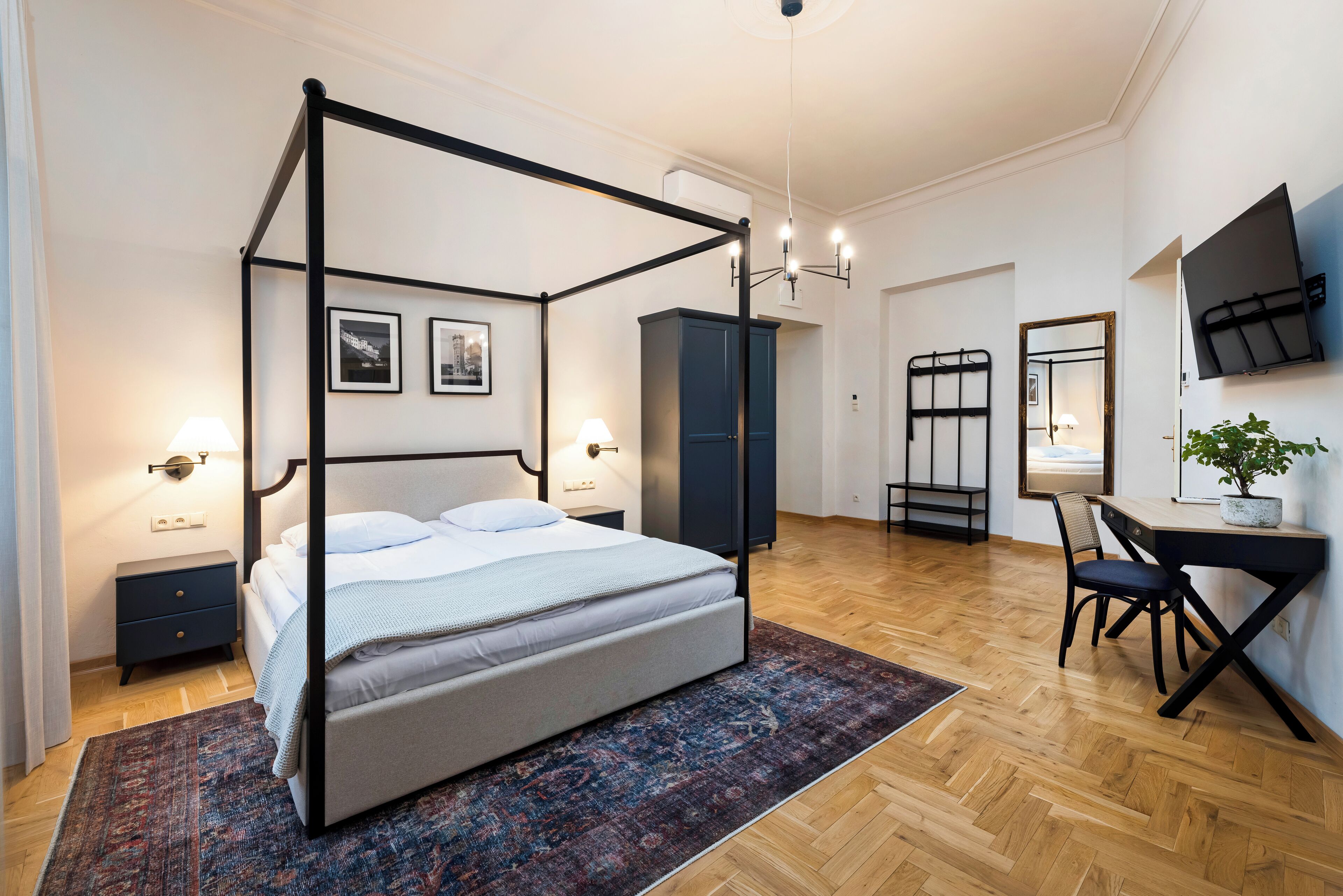A modern bedroom with a four-poster bed, a desk with a chair, a television mounted on the wall, and a wardrobe.