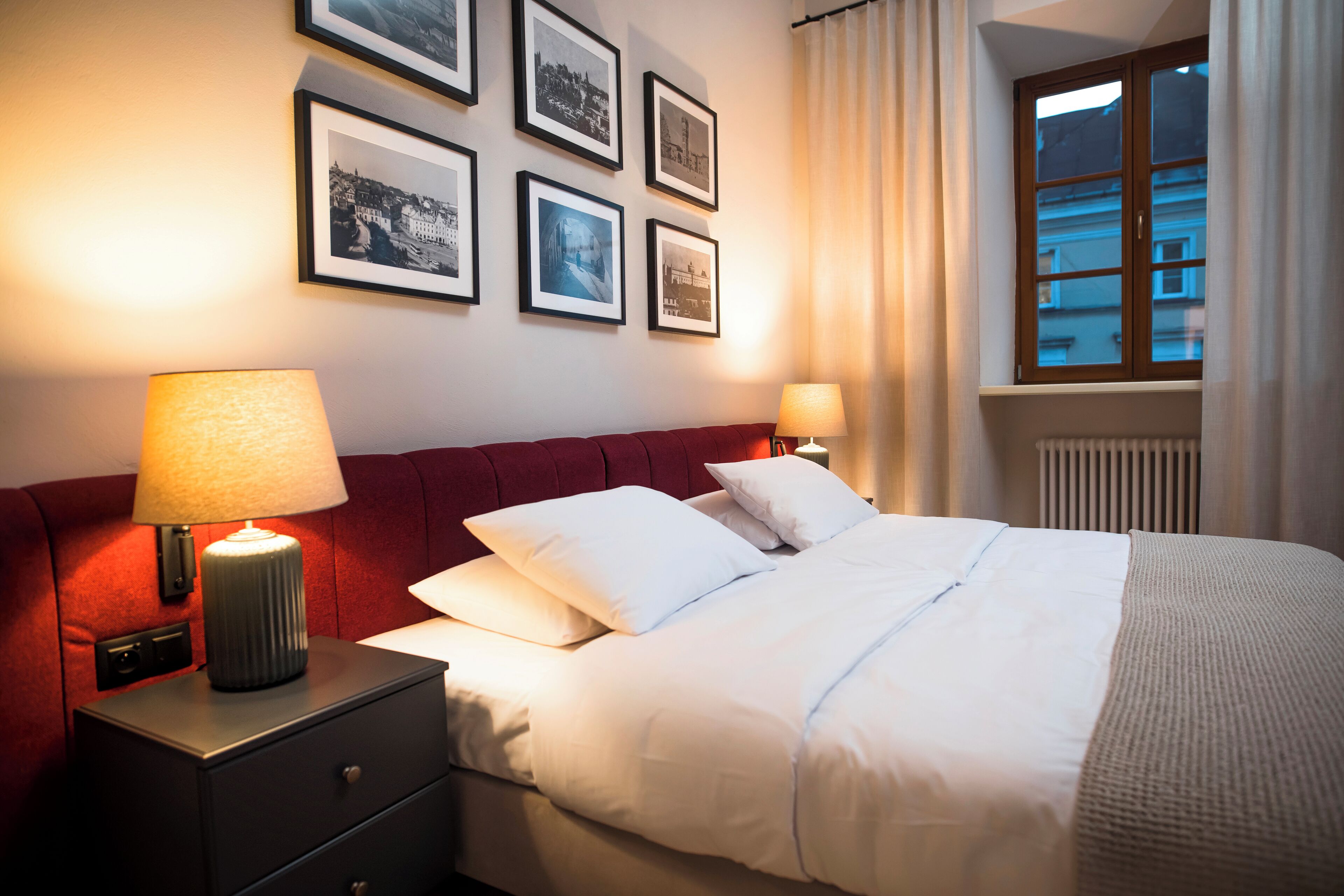 Hotellihuone, jossa on sänky, kaksi yöpöytälamppua, yöpöytä ja ikkuna, jossa on verhot.