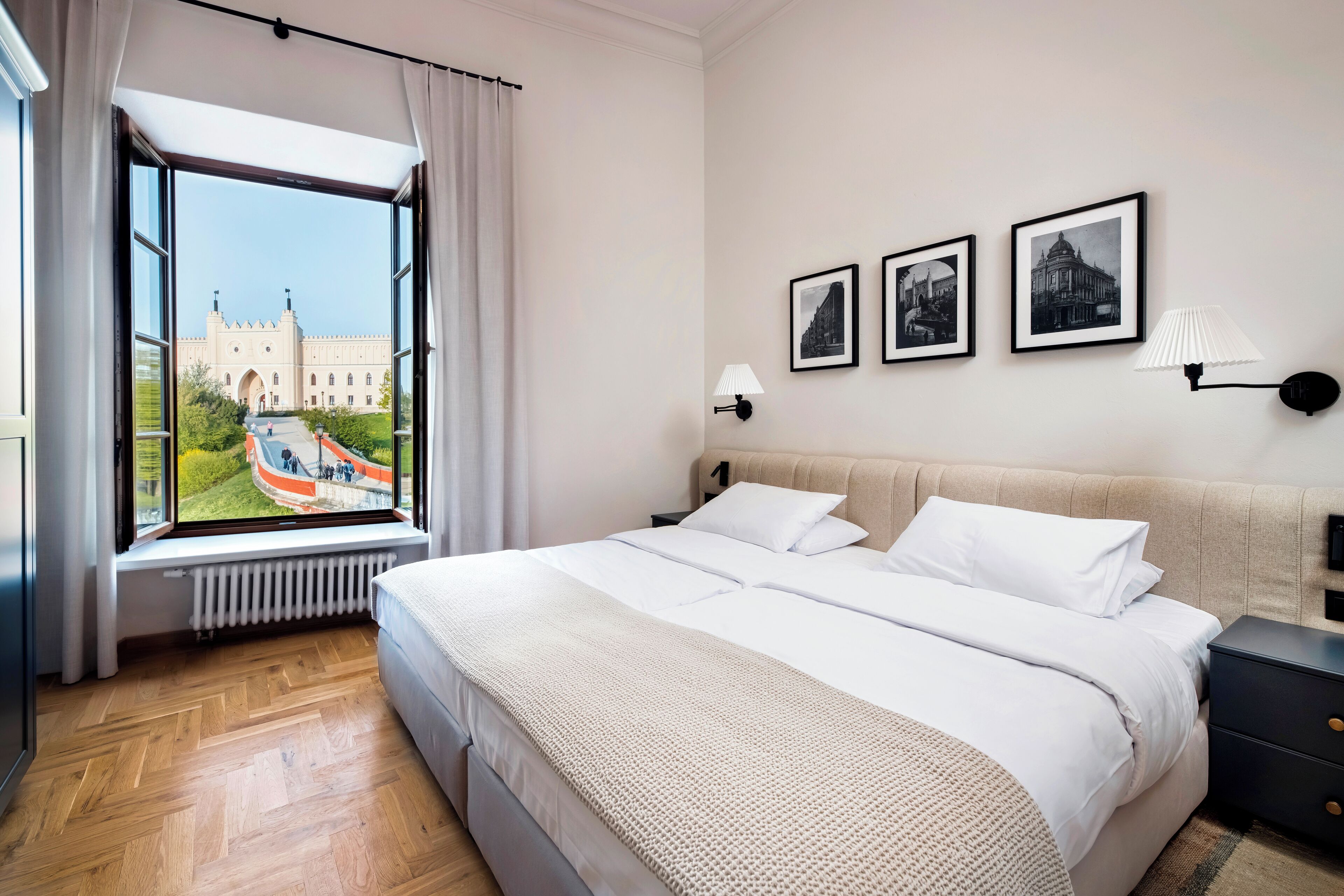 A hotel room with a large bed, a window with a view of a castle, and three framed pictures on the wall.