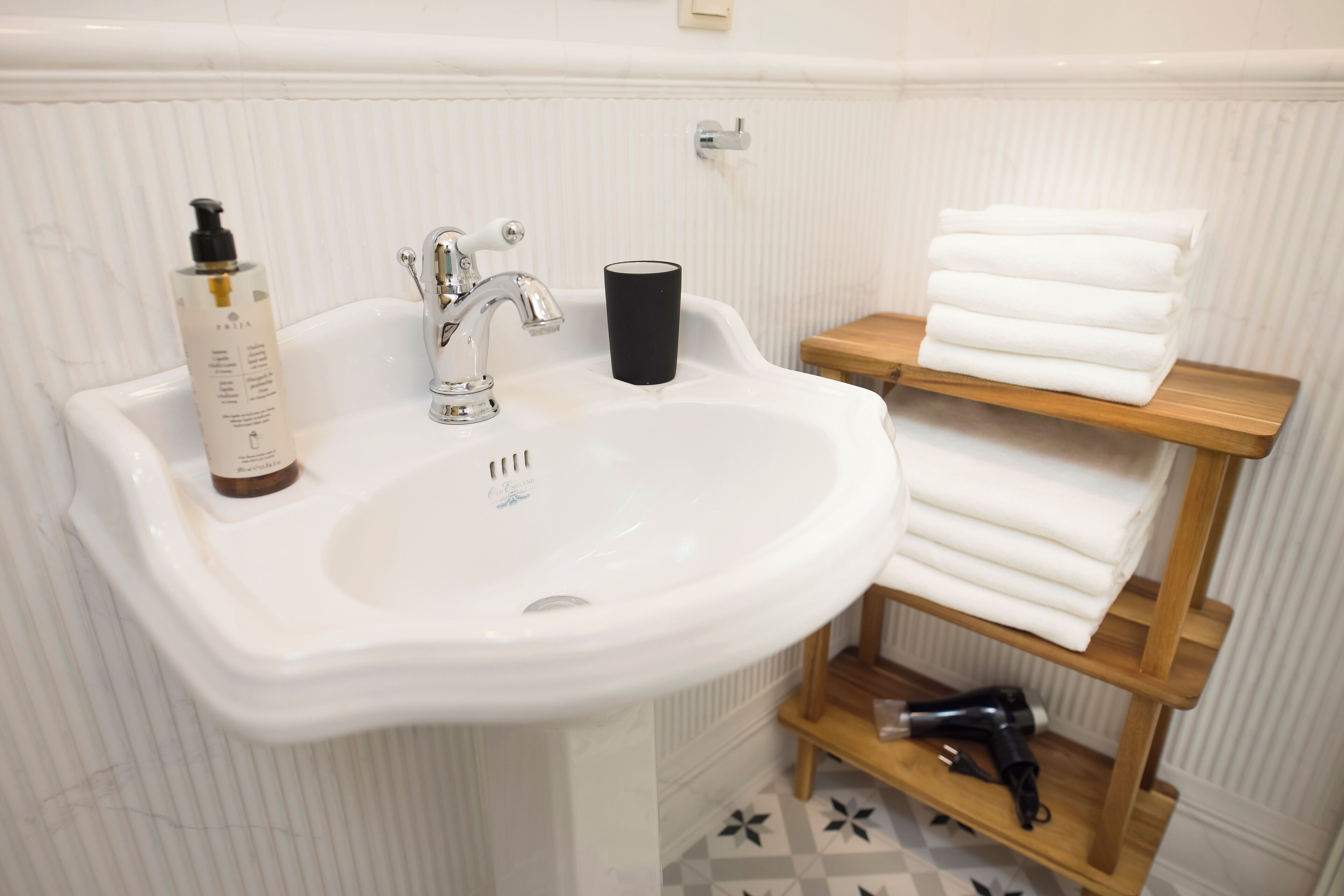 A bathroom with a white sink, a black cup, a bottle of shampoo, and a shelf with folded towels.