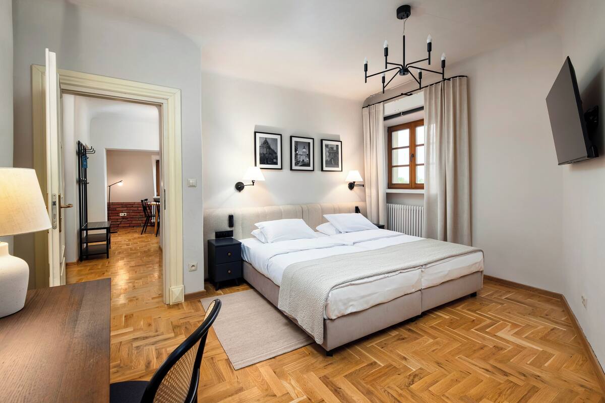 Une chambre à coucher avec un lit, des tables de chevet, un bureau et une chaise. Les murs sont ornés de tableaux encadrés, il y a une fenêtre avec des rideaux et un luminaire au plafond.