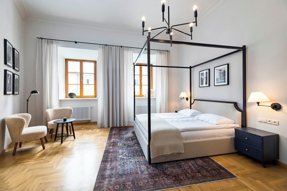 Un lit à baldaquin avec un cadre sombre, une table de chevet avec une lampe et une chaise dans une pièce dotée d’un plancher en bois et d’un tapis à motifs.