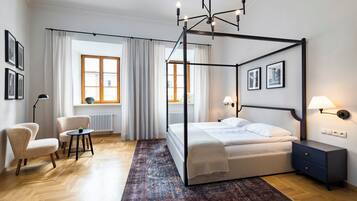A four-poster bed with a dark frame, a bedside table with a lamp, and a chair in a room with wooden flooring and a patterned rug.