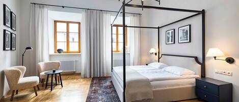 Una cama con dosel de estructura oscura, una mesita de noche con lámpara y una silla en una habitación con suelo de madera y alfombra estampada.