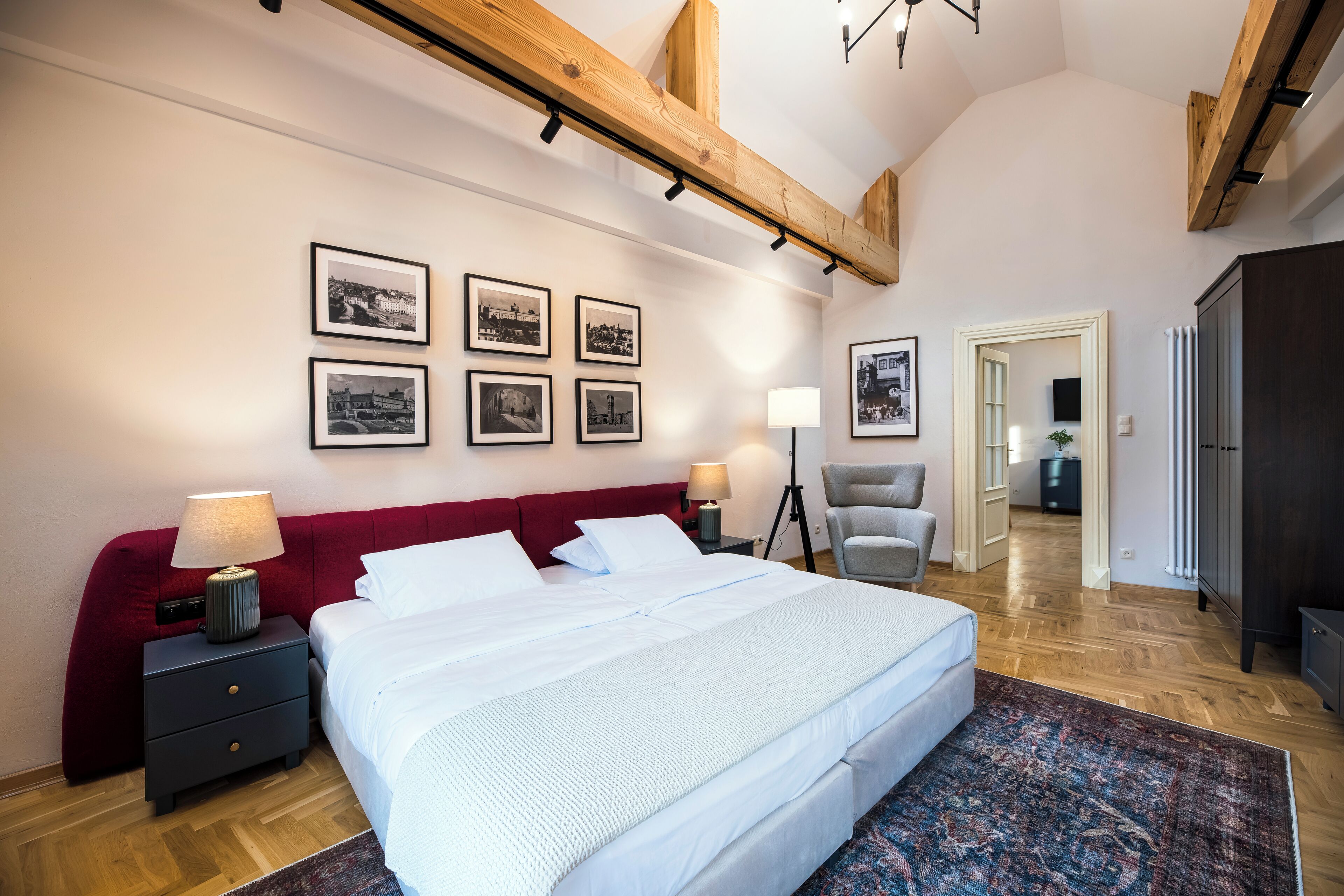A bedroom with a large bed, a red headboard, a nightstand with a lamp, and a chair. There are framed pictures on the wall and a hallway leading to another room.