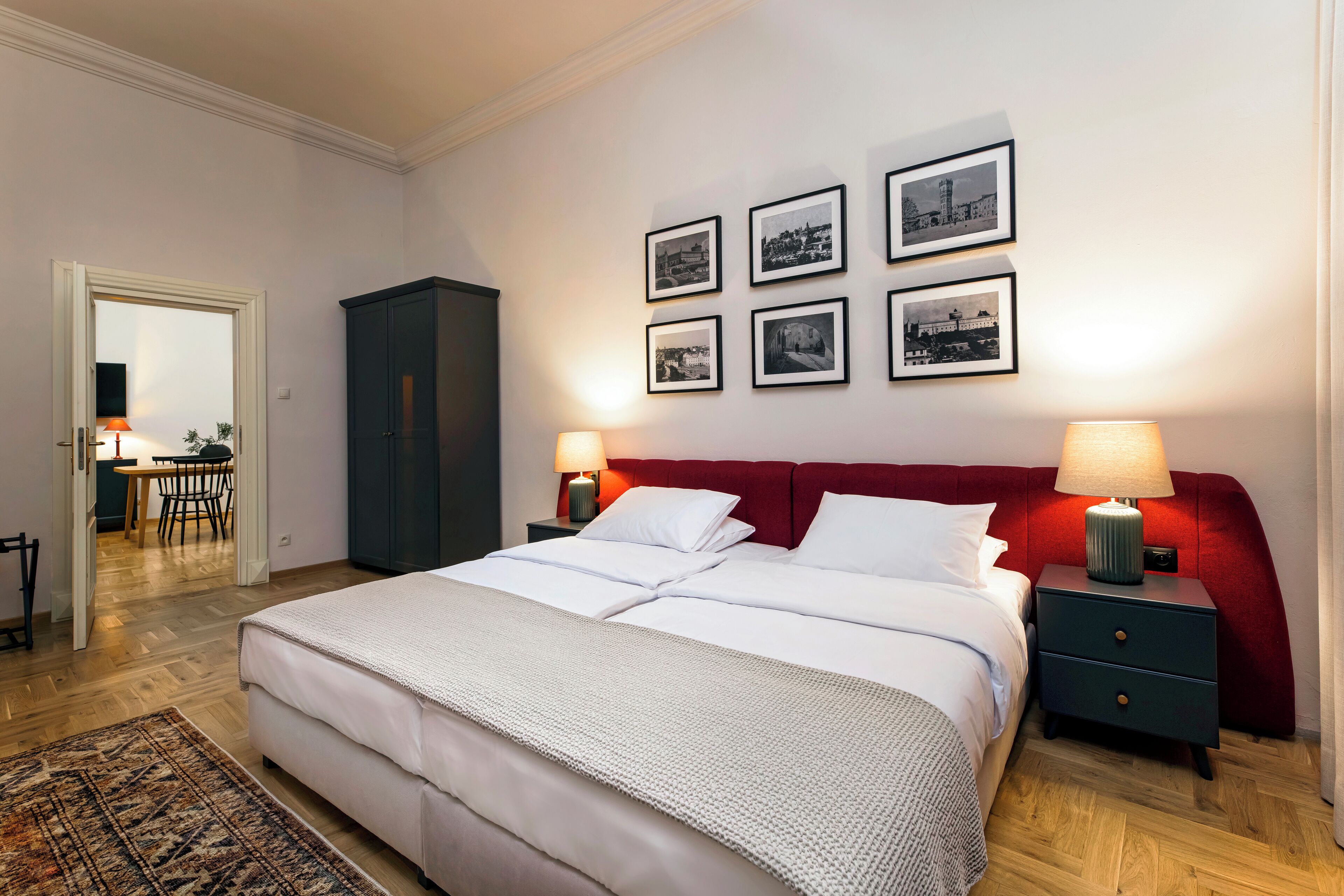 Un dormitorio ordenado con una cama grande, dos mesitas de noche con lámparas, un cabecero rojo y seis fotografías blanco y negro en la pared.