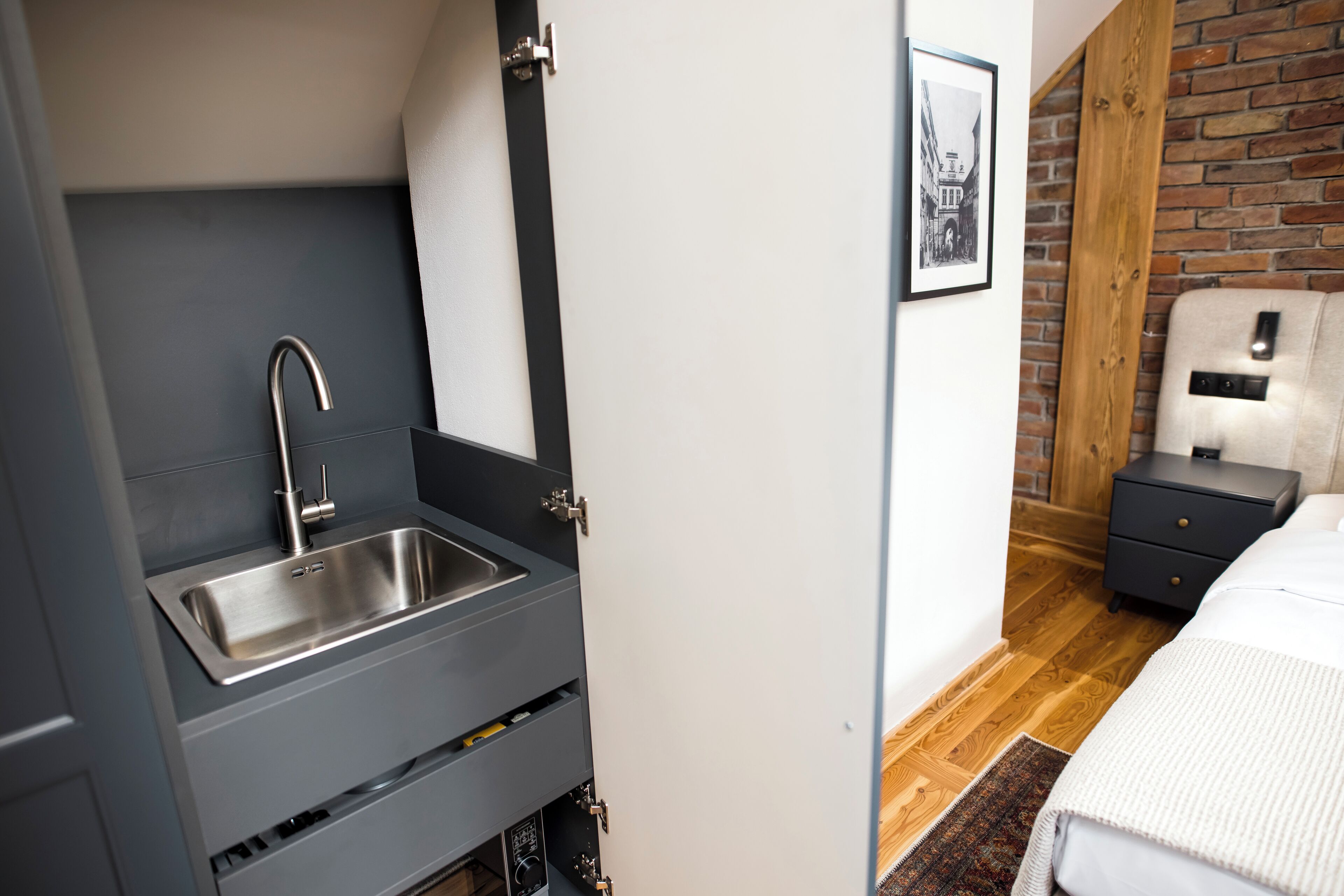 A compact room with a built-in sink, a bed with a white comforter, a nightstand, and a brick-patterned wall.