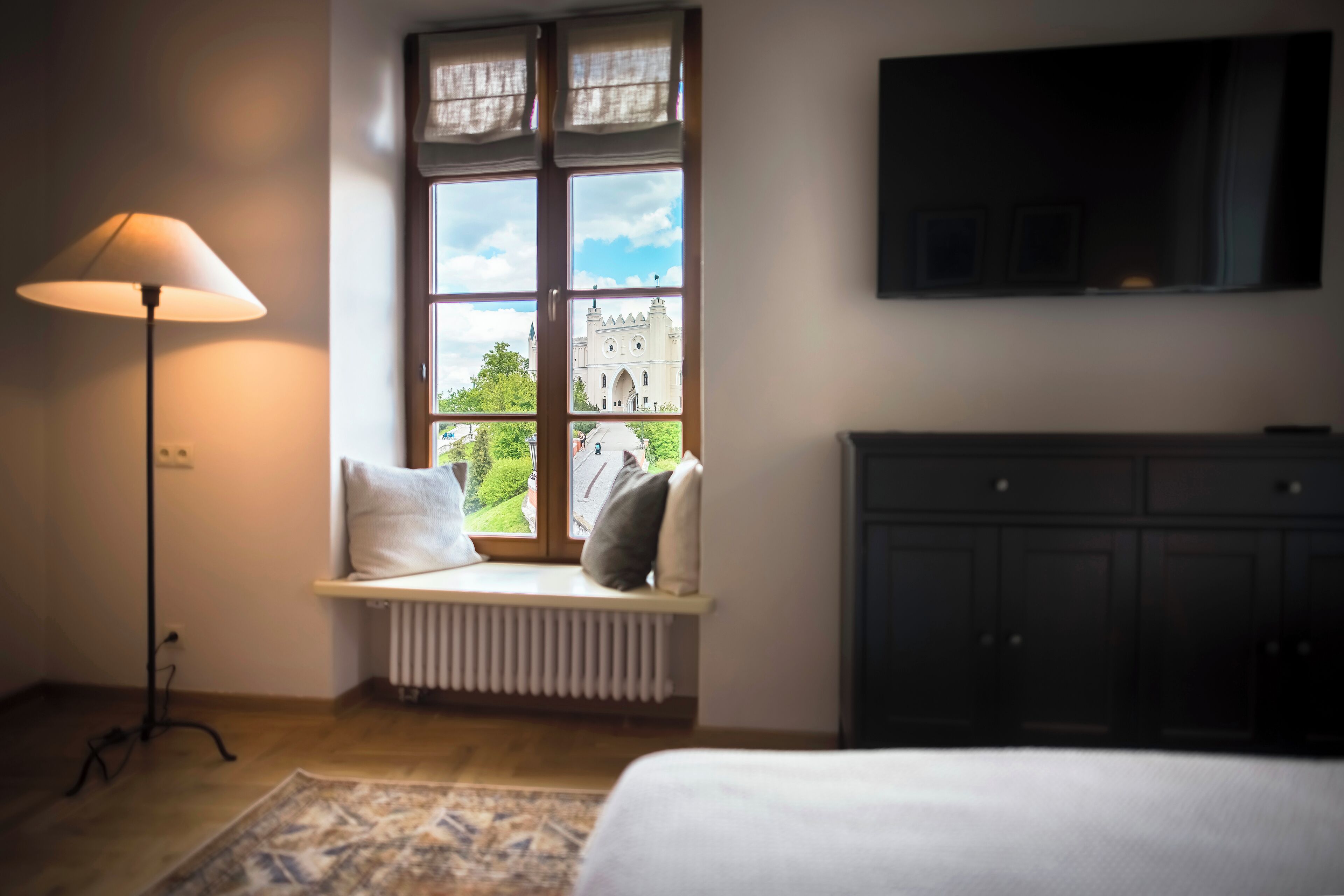 A bedroom with a large window, a radiator, a floor lamp, a bed, and a TV mounted on the wall.