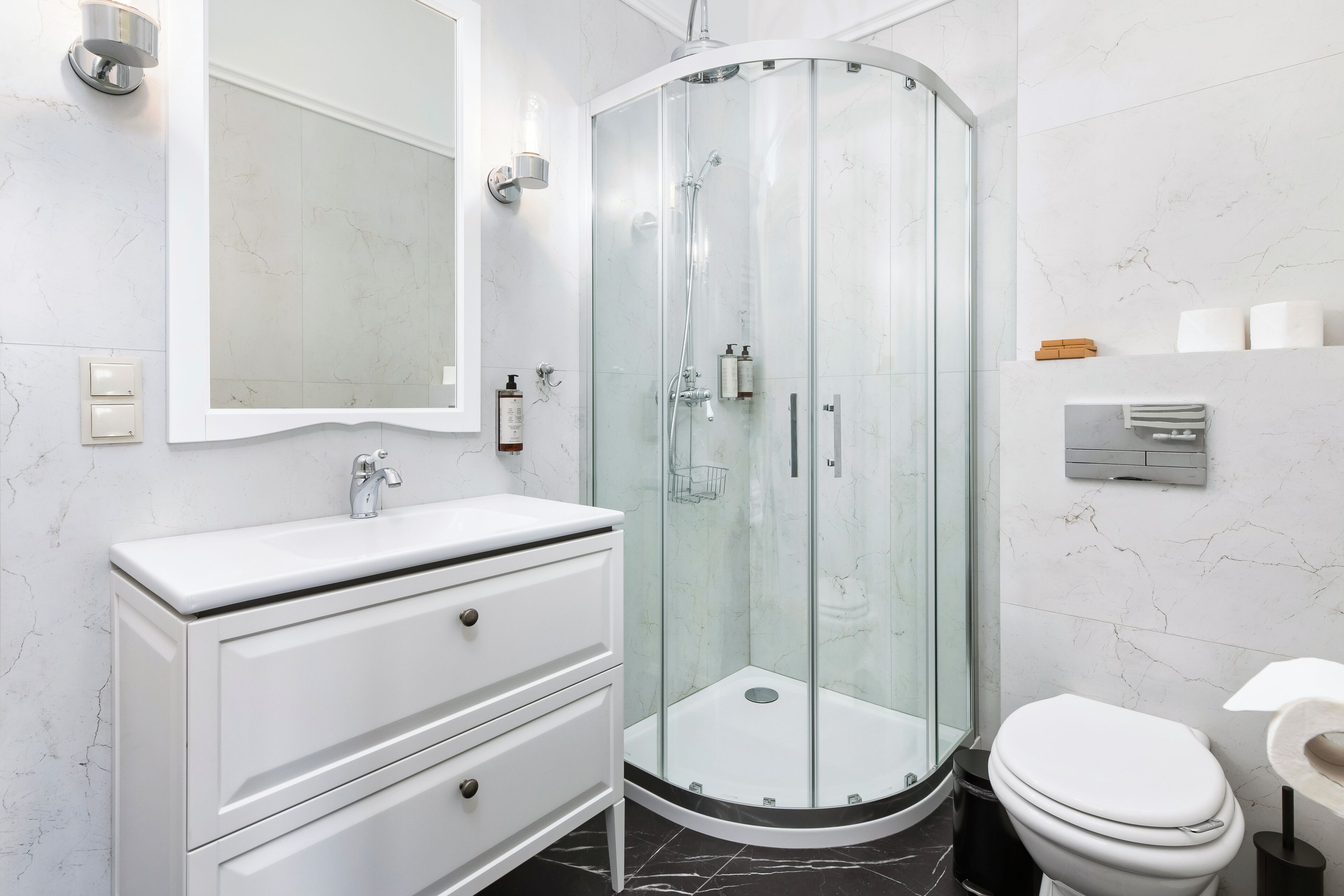 Un baño moderno con cabina de ducha de vidrio, tocador blanco con cajones, inodoro y paredes de mármol.