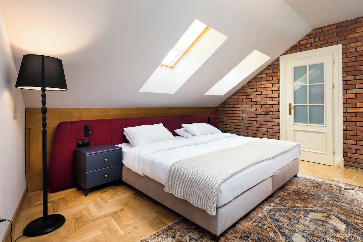 Une chambre à coucher avec un lit, une table de chevet, une lampe, une tête de lit rouge, un mur en briques et des fenêtres de toit.