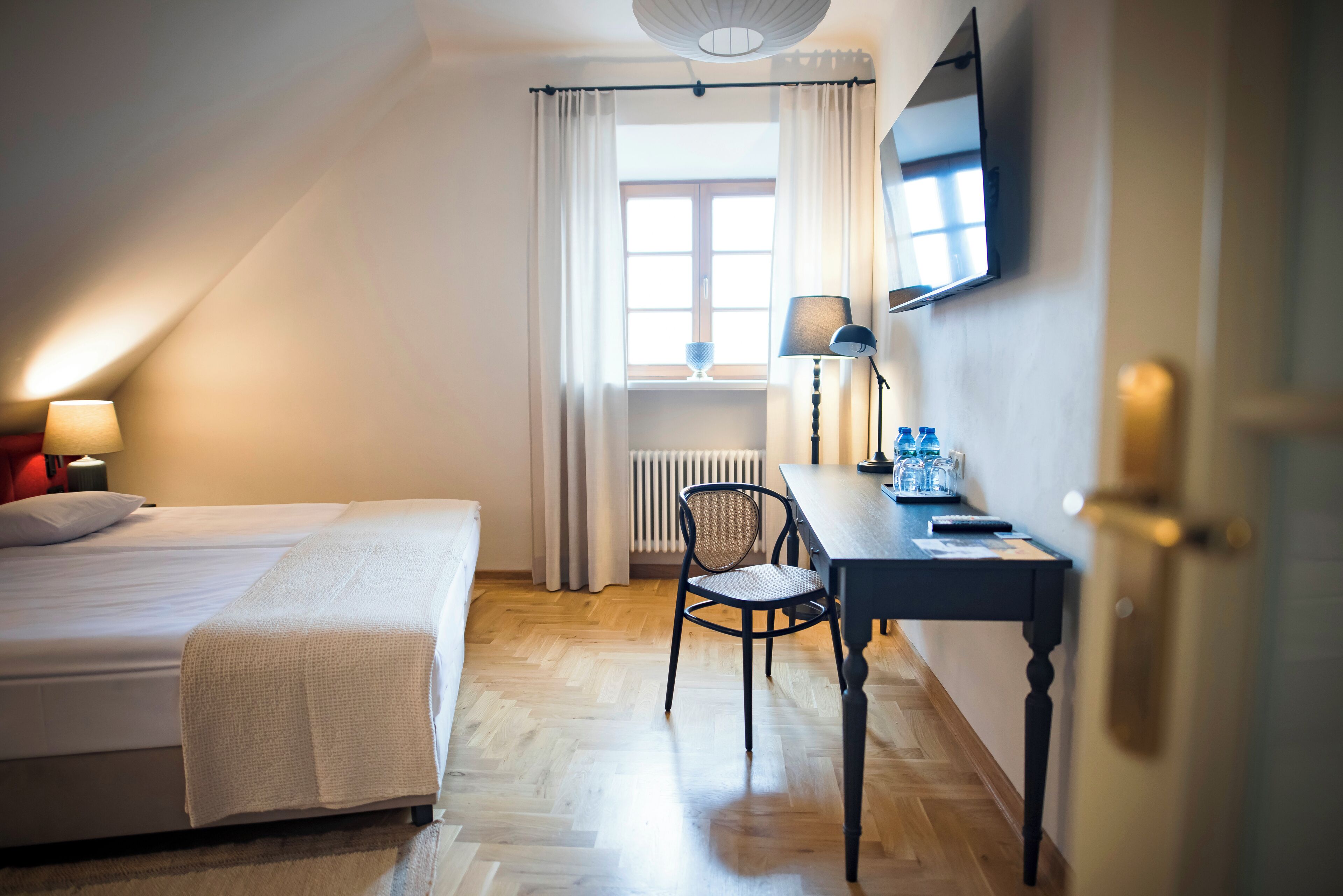 A hotel room with a bed, a desk, a chair, a TV, and a window with curtains.