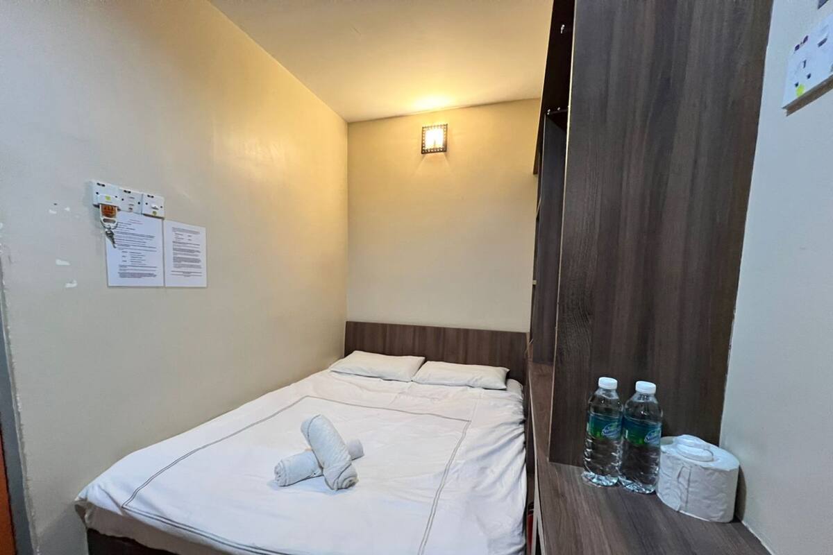 A small, neatly made bed with white linens and a rolled towel, a wooden headboard, a wall-mounted light, and a small table with bottled water and toilet paper.
