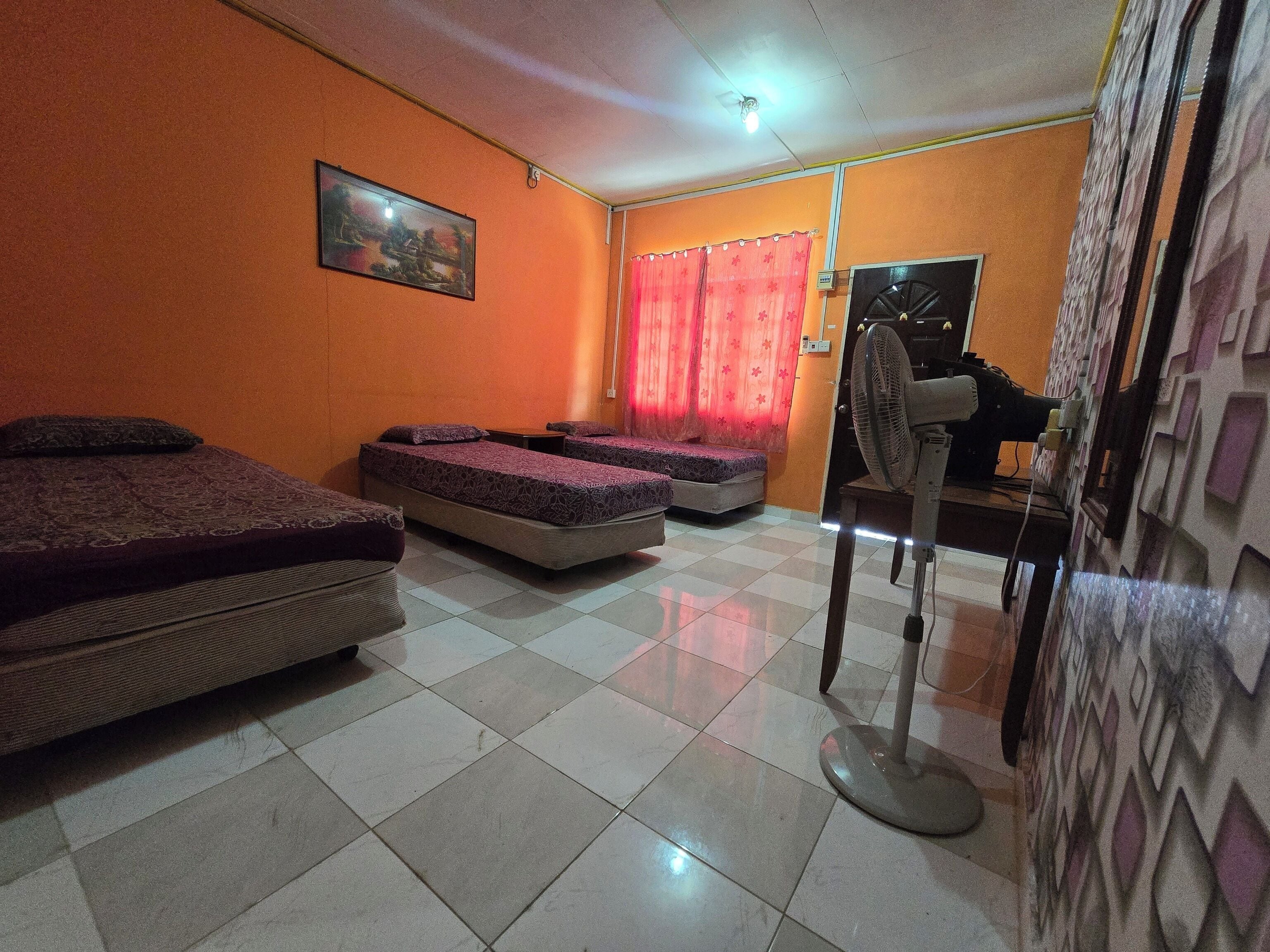A room with two beds, a standing fan, and a window with red curtains.