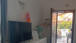 Een kamer met een flatscreen-tv op een witte kast, een ventilator en een deur die leidt naar een buitenruimte met betegelde vloer.