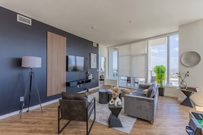 A modern living room with a large window, a flat-screen TV, and a city view. - Modern Skyline Retreat | Pool|Gym, Balcony&Parking (Phoenix)