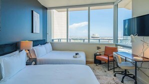 A hotel room with two beds, a desk, a chair, a TV, and a large window with a city view.