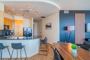 Una cocina moderna con una isla central, taburetes de bar, una mesa de comedor y una sala de estar con televisor y lámpara de pie.