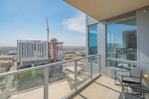 A balcony with a glass railing, a chair, and a view of a cityscape including a construction crane. - Game Day in PHX | Pool,Gym,Parking,Balcony,W/D (Phoenix)
